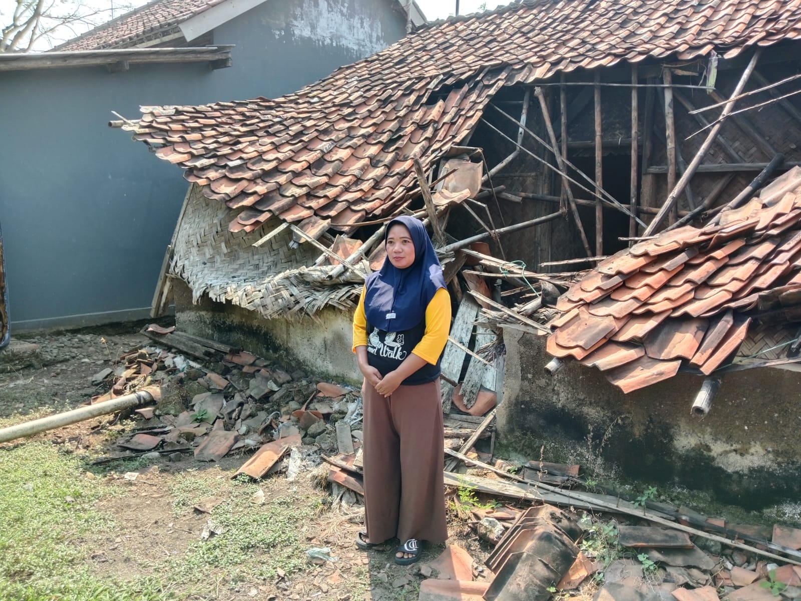Caption : Ibu Sarwi berdiri di depan rumahnya yang roboh di Dusun Jatiboros RT 15/RW 05, Desa Kertajaya, Kecamatan Jayakerta, Kabupaten Karawang. Enam bulan pascakejadian akibat hujan deras disertai angin kencang, keluarga masih terpaksa menumpang di rumah kerabat sambil berharap adanya bantuan renovasi dari Pemerintah Kabupaten Karawang. (Foto: Gugun/Patriotjabar.com)