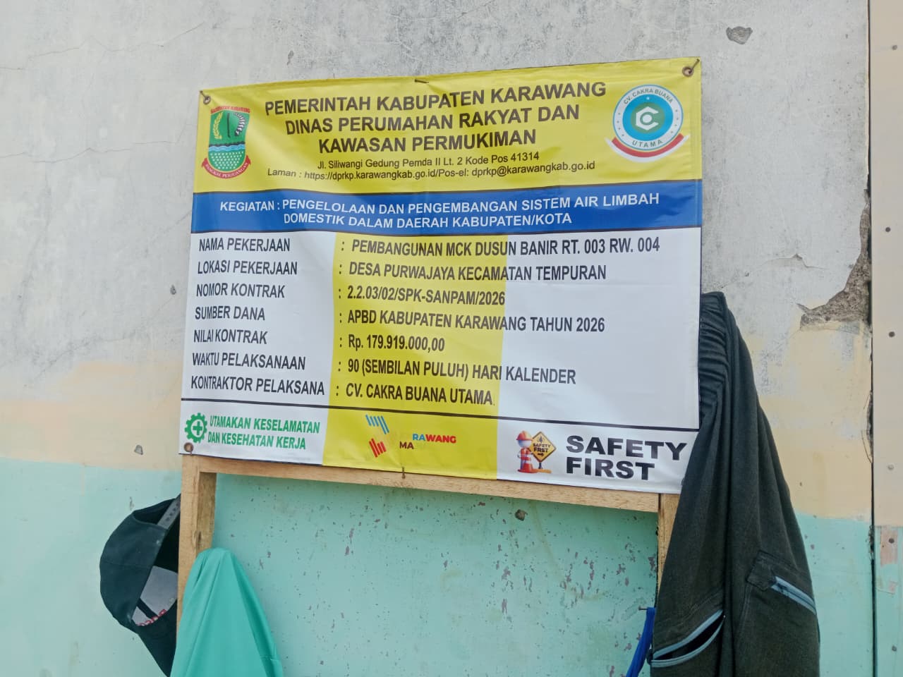 Caption: Papan informasi proyek pembangunan MCK Dusun Banir, Desa Purwajaya, Kecamatan Tempuran, Kabupaten Karawang, dengan nilai kontrak Rp179,919 juta yang bersumber dari APBD Kabupaten Karawang Tahun Anggaran 2026 dan dikerjakan CV.Cakra Buana Utama.