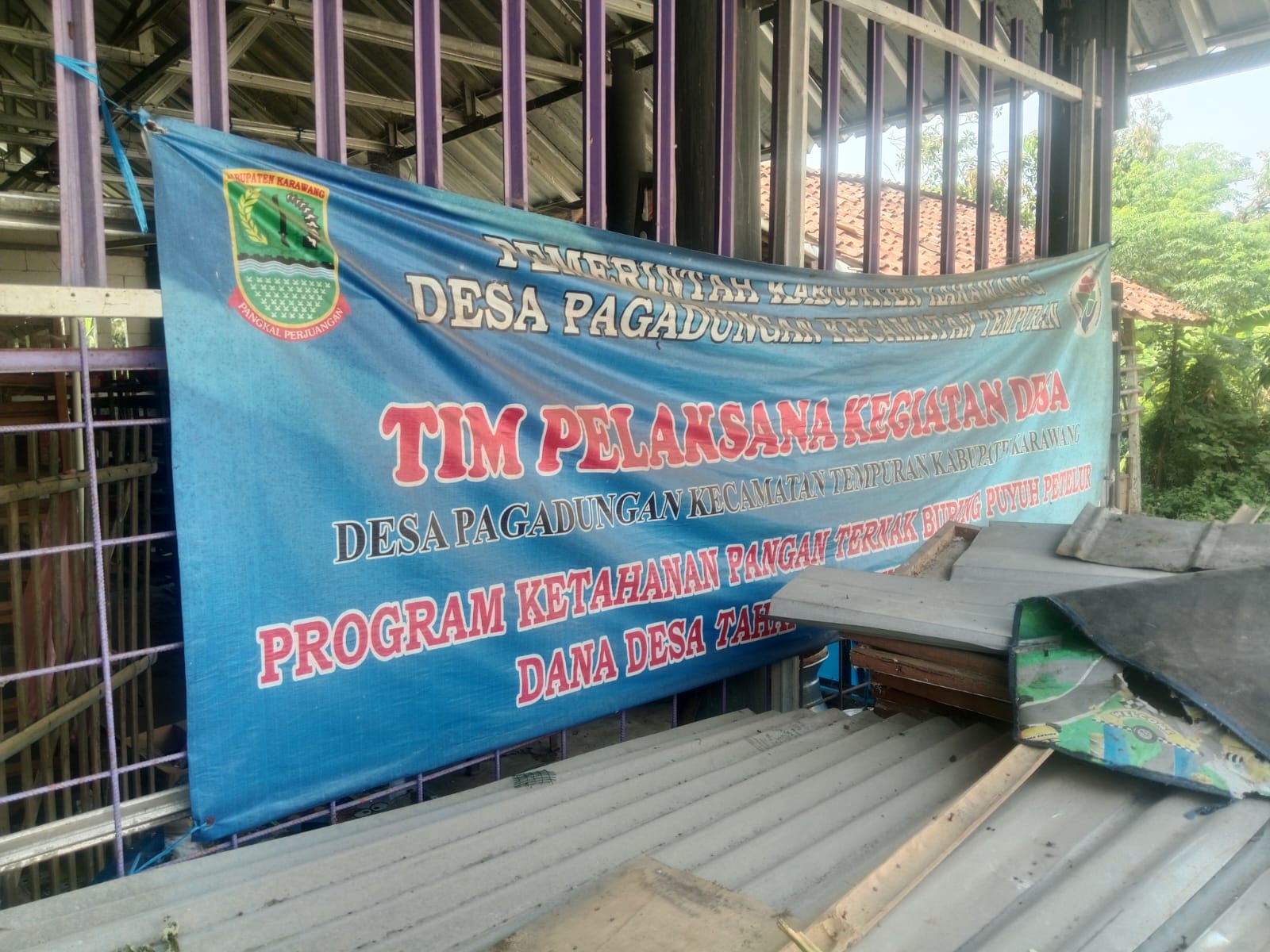 Caption: Spanduk program ketahanan pangan berupa budidaya burung puyuh dan jangkrik yang bersumber dari Dana Desa Tahap II Tahun Anggaran 2025 di Desa Pegadungan, Kecamatan Tempuran, Kabupaten Karawang. Kondisi program tersebut kini menjadi sorotan setelah aktivitas peternakan diduga berhenti.