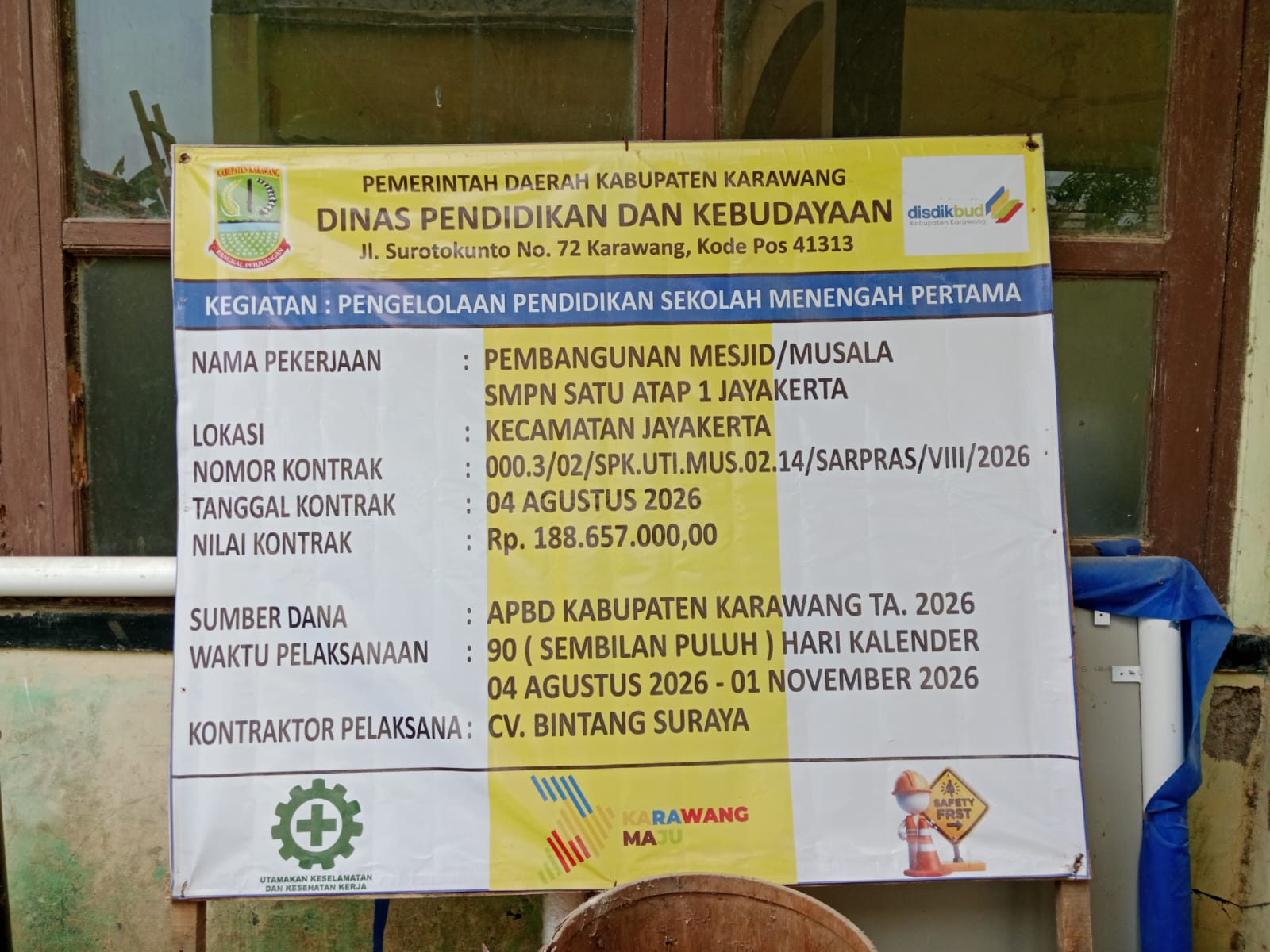 Caption: Papan informasi proyek pembangunan Masjid/Musala SMPN Satu Atap 1 Jayakerta, Kabupaten Karawang. Proyek bersumber dari APBD Kabupaten Karawang TA 2026 dengan nilai kontrak Rp188.657.000, dikerjakan CV Bintang Suraya selama 90 hari kalender, terhitung 4 Agustus hingga 1 November 2026.